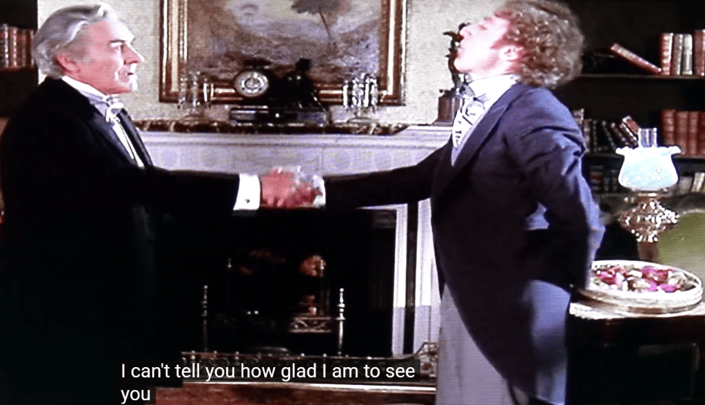 Two men in formal attire are shaking hands in a vintage living room setting, with a fireplace and bookshelves in the background. One man expresses happiness to see the other.
