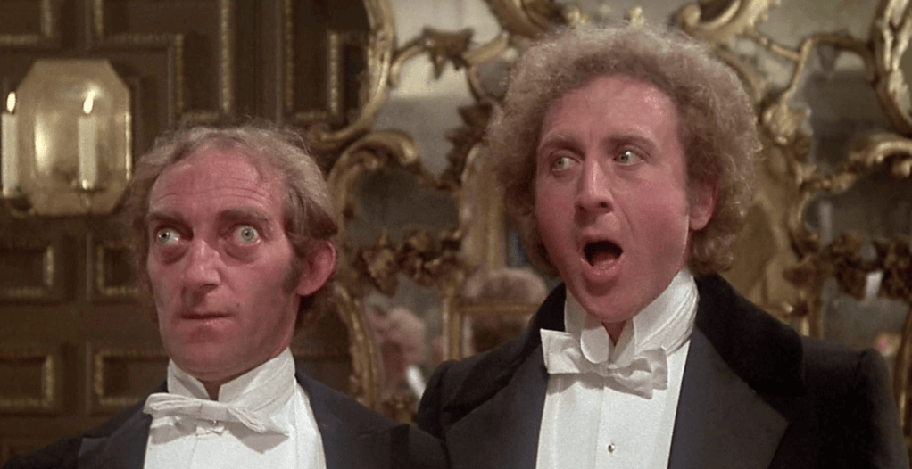 Two men in tuxedos with exaggerated facial expressions, one looking surprised and the other appearing to be singing or shouting, set against an ornate background with mirrors.