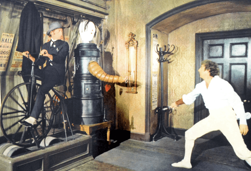 A scene featuring a man on a bicycle-powered contraption with a large mechanical arm in a vintage setting, while another man in a white outfit appears to confront him.