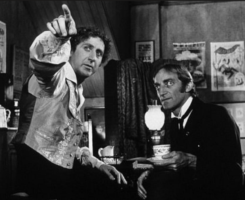 Black and white image of two men in period clothing, one pointing and gesturing while the other looks on attentively, both seated at a table with a cup in front of them. A lantern and posters are visible in the background.