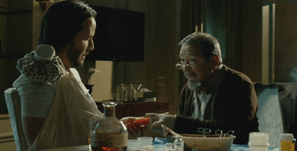 A man in a robe sits in a dimly lit room, receiving care from an elderly man wearing glasses and a cardigan, who is holding medical items.