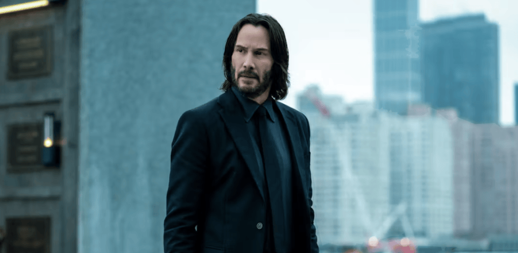 A man in a black suit stands outdoors, looking thoughtfully towards the distance, with a city skyline in the background.